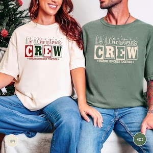 以下が含まれることがあります： 「Christmas CREW」の文字がデザインされた、アイボリーとモスグリーンの2枚のTシャツ。下には「MAKING MEMORIES TOGETHER」の文字。アイボリーのシャツはブルージーンズと組み合わせています。モスグリーンのシャツもブルージーンズと組み合わせています。