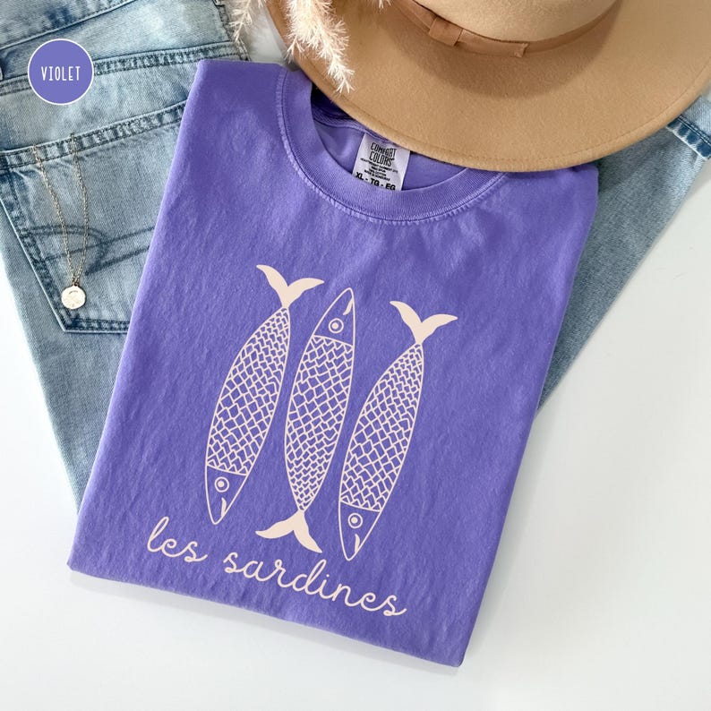 Op de afbeelding: Een paars T-shirt met een afbeelding van drie gestileerde vissen en de tekst "les sardines".
