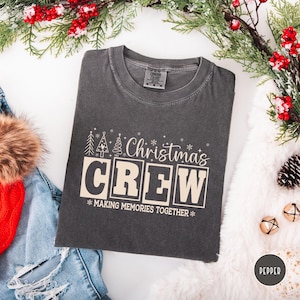 以下が含まれることがあります： クリーム色の「Christmas CREW」の文字が入った濃いグレーのTシャツ。デザインにはクリスマスツリーと雪の結晶のグラフィックが含まれています。「MAKING MEMORIES TOGETHER」のフレーズが「CREW」の下に印刷されています。