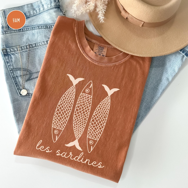 Op de afbeelding: Een roestkleurig T-shirt met een afbeelding van drie gestileerde vissen en de tekst "les sardines".