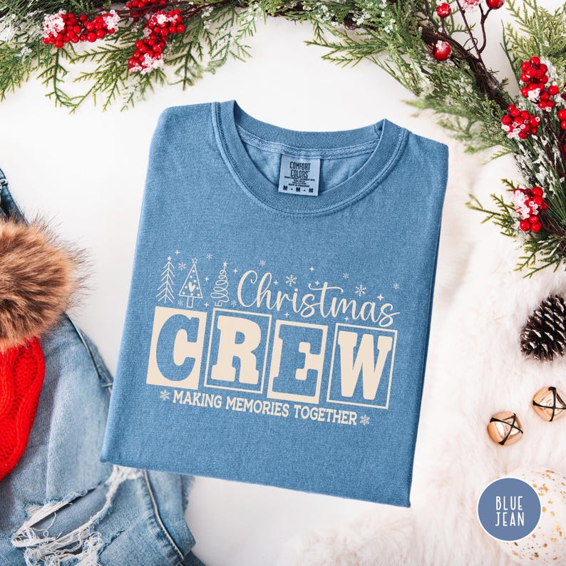 以下が含まれることがあります： クリーム色の「Christmas CREW」の文字が入った水色のTシャツ。Tシャツには「MAKING MEMORIES TOGETHER」というフレーズとクリスマスツリーのグラフィックも入っています。Tシャツはクリスマスの飾りで囲まれています。