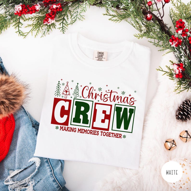 以下が含まれることがあります： 赤と緑の「Christmas CREW」の文字とクリスマスツリーのグラフィックがプリントされた白いTシャツ。「MAKING MEMORIES TOGETHER」のフレーズが「CREW」の下にあります。Tシャツはクリスマスの装飾で囲まれています。