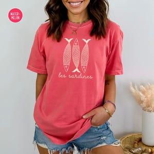 Op de afbeelding: Een koraalroze T-shirt met een afbeelding van drie witte vissen en de tekst "les sardines" in wit.