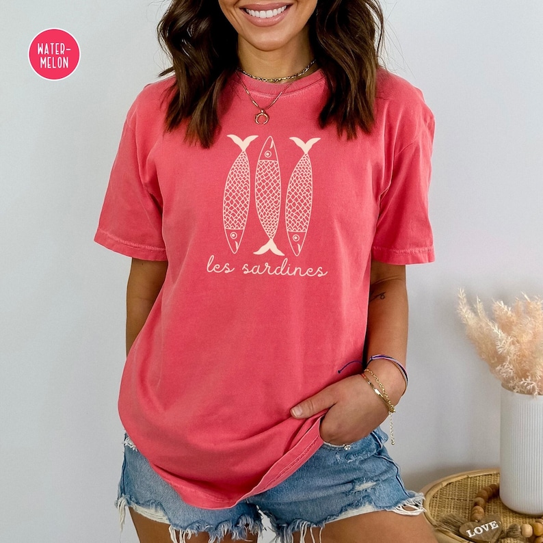Op de afbeelding: Een koraalroze T-shirt met een afbeelding van drie witte vissen en de tekst "les sardines" in wit.