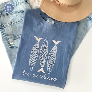 Op de afbeelding: Een blauw T-shirt met een witte afbeelding van drie gestileerde vissen en de tekst "les sardines".
