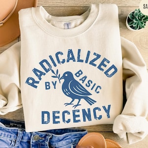 Op de afbeelding: Een crèmekleurige sweatshirt met de tekst "RADICALIZED BY BASIC DECENCY" in verweerde blauwe letters. Een blauwe duif-afbeelding houdt een olijftak vast. De sweatshirt is op een houten oppervlak gevouwen.