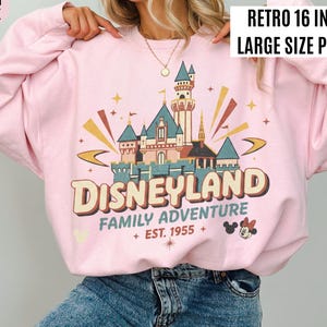 May include: Light pink sweatshirt with a retro Disneyland graphic. The design features a castle, the word "Disneyland", and the text "Family Adventure Est. 1955". The print is 16 inches in size.