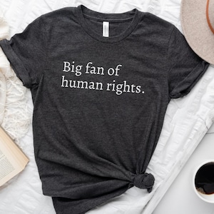 May include: Dark gray t-shirt with the text "Big fan of human rights" in white. The shirt is knotted at the bottom. A beige hat, a cup of coffee, and a book are in the background.