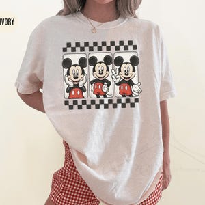 Puede incluir: Camiseta de color marfil con un gr&aacute;fico de Mickey Mouse en tres poses diferentes, sobre un fondo a cuadros blanco y negro. Mickey lleva pantalones cortos rojos y guantes blancos. La camiseta se combina con pantalones cortos a cuadros rojos y blancos.