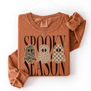 May include: A long-sleeve crewneck sweatshirt in a burnt orange colour. The front features the black text "SPOOKY SEASON" and three ghost illustrations with different patterns: camouflage, leopard print, and a brown and tan checkerboard design.