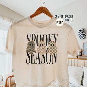 May include: An ivory boxy t-shirt featuring the black text SPOOKY SEASON and three ghost illustrations. The ghosts have different patterns: one with camouflage, one with leopard print, and one with a tan and white checkerboard pattern.