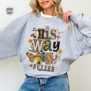 Może przedstawiać: Szara bluza sportowa z napisem "His Way Is Better" w kolażu czcionek i obrazów, w tym motyla, kwiatów i krzyża. Bluza jest w kolorze jasnoszarym.
