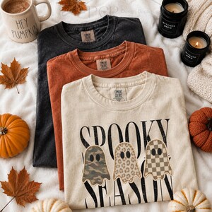 May include: Three crewneck t-shirts in black, burnt orange, and cream. The cream shirt features the black text SPOOKY SEASON and three ghost graphics with camouflage, leopard print, and checkerboard patterns. Each shirt has a Comfort Colors label.
