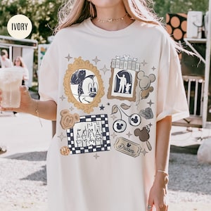 May include: Ivory-colored t-shirt featuring a collage of Disney-themed graphics. The design includes a Mickey Mouse portrait, a silhouette of a person with a dog, and various other elements like cherries and a cookie. The word "Dream" is also visible.