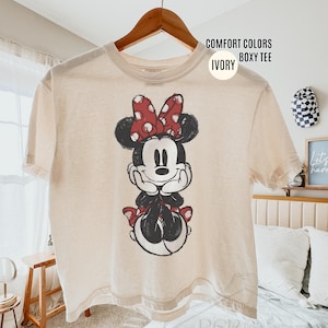 May include: Ivory-colored boxy tee shirt featuring a graphic of Minnie Mouse. Minnie is depicted in black and white with a red polka-dot bow and accents. The shirt is hanging on a wooden hanger.