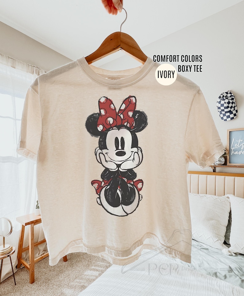 May include: Ivory-colored boxy tee shirt featuring a graphic of Minnie Mouse. Minnie is depicted in black and white with a red polka-dot bow and accents. The shirt is hanging on a wooden hanger.