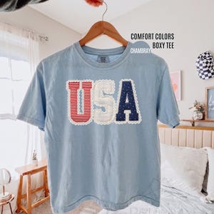 May include: A light blue short-sleeve boxy t-shirt featuring the text USA in large applique letters. The U is red and white striped, the S is white, and the A is navy blue with white dots. Each letter is outlined with white lace trim.