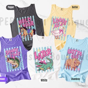 K&ouml;nnte beinhalten: Eine Sammlung von sechs bunten Tanktops mit verspielten Grafiken und Texten. Designs umfassen ein Pferd, eine Ente, einen Fisch, ein Alligator und einen B&uuml;ffel, mit S&auml;tzen wie "Hold Your", "Loose As A" und "Gotta Go". Die Tanktops sind in verschiedenen Farben.