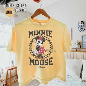 May include: Yellow boxy tee shirt with a Minnie Mouse graphic. The graphic features Minnie Mouse in a circle with a checkered background, the words "MINNIE MOUSE" and the Disney logo. The shirt is hanging on a wooden hanger.