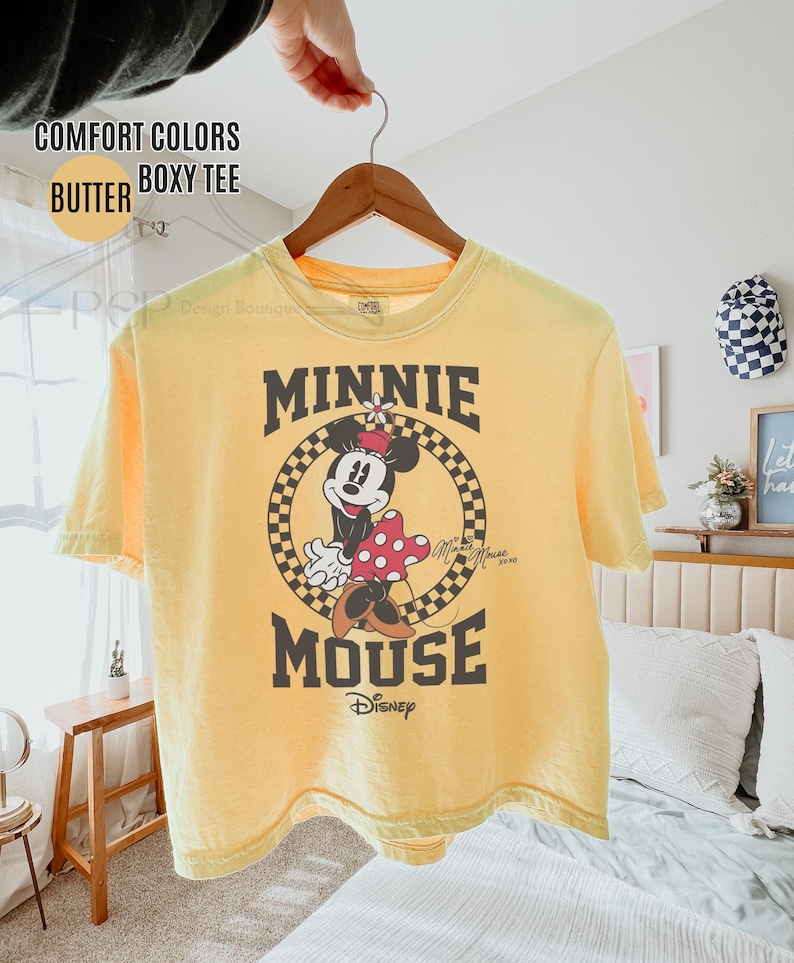 May include: Yellow boxy tee shirt with a Minnie Mouse graphic. The graphic features Minnie Mouse in a circle with a checkered background, the words "MINNIE MOUSE" and the Disney logo. The shirt is hanging on a wooden hanger.