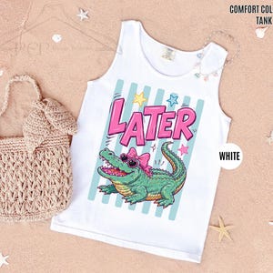 K&ouml;nnte beinhalten: Wei&szlig;es Tanktop mit einem Cartoon-Alligator, der eine Sonnenbrille und eine rosa Schleife tr&auml;gt. Das Wort "LATER" steht in rosa Buchstaben. Das Tanktop ist mit "COMFORT COLORS TANK TOP" und "WHITE" beschriftet.