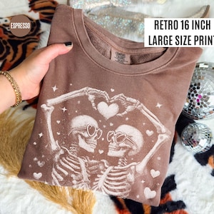 May include: Espresso-colored sweatshirt with a retro 16-inch large-size print. The design features two skeletons forming a heart with their arms, wearing heart-shaped sunglasses. White hearts and stars are scattered around the skeletons.