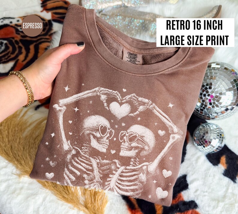 May include: Espresso-colored sweatshirt with a retro 16-inch large-size print. The design features two skeletons forming a heart with their arms, wearing heart-shaped sunglasses. White hearts and stars are scattered around the skeletons.