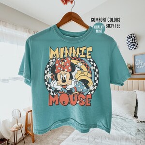 Może przedstawiać: Miętowa koszulka Comfort Colors z nadrukiem Minnie Mouse. Projekt zawiera twarz Minnie, czerwoną kokardę i napis "MINNIE MOUSE" w stylu retro. Koszulka wisi na drewnianym wieszaku.