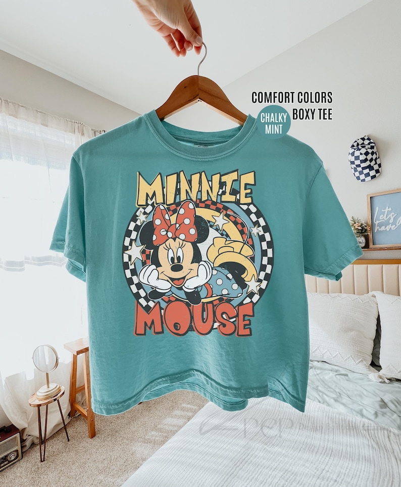Może przedstawiać: Miętowa koszulka Comfort Colors z nadrukiem Minnie Mouse. Projekt zawiera twarz Minnie, czerwoną kokardę i napis "MINNIE MOUSE" w stylu retro. Koszulka wisi na drewnianym wieszaku.