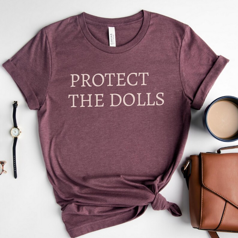 Puede incluir: Una camiseta de color granate con el texto "PROTECT THE DOLLS" impreso en blanco.