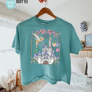 May include: Mint green boxy tee featuring a colorful graphic of a fairytale castle, fireworks, and Tinkerbell. The shirt has the words "COMFORT COLORS", "CHALKY", "BOXY TEE", and "MINT" printed on the top left.