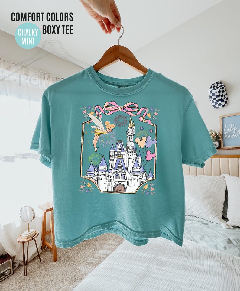 May include: Mint green boxy tee featuring a colorful graphic of a fairytale castle, fireworks, and Tinkerbell. The shirt has the words "COMFORT COLORS", "CHALKY", "BOXY TEE", and "MINT" printed on the top left.