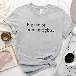May include: A light gray t-shirt with the text "Big fan of human rights." The shirt is knotted at the bottom and is surrounded by a book, a candle, a sleep mask, a hat, and a cup of coffee.