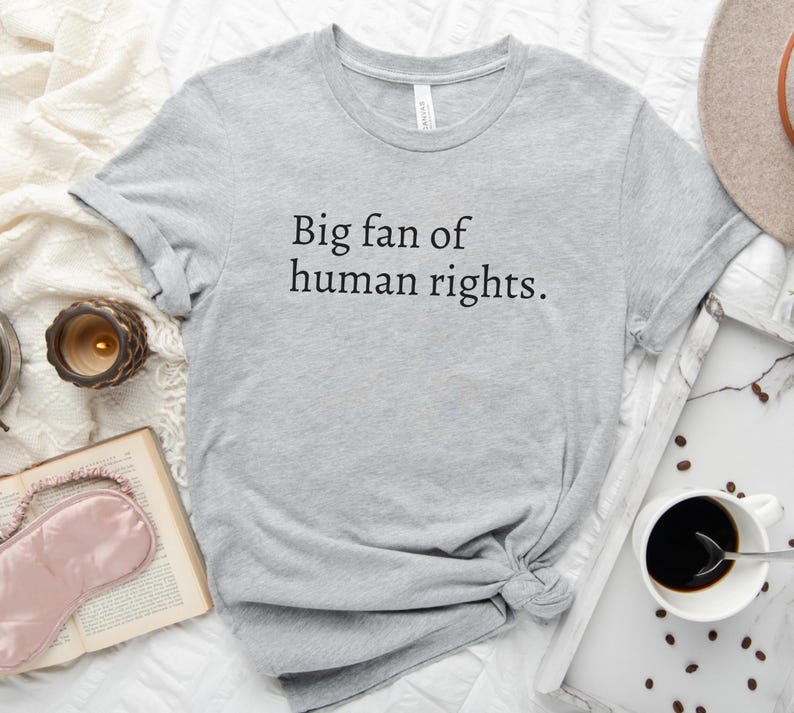 May include: A light gray t-shirt with the text "Big fan of human rights." The shirt is knotted at the bottom and is surrounded by a book, a candle, a sleep mask, a hat, and a cup of coffee.
