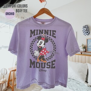May include: Orchid-colored boxy tee featuring a graphic of Minnie Mouse. The design includes Minnie in a polka-dot dress, set within a checkered circle, with the words "MINNIE MOUSE" above and below. The Disney logo is at the bottom.