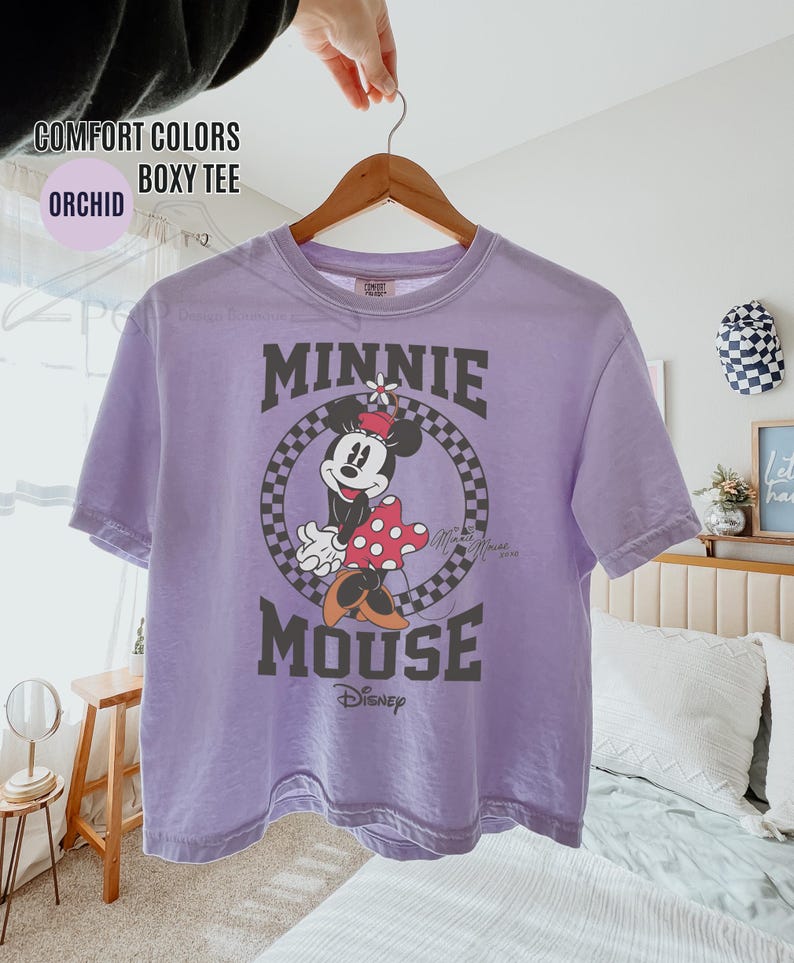 May include: Orchid-colored boxy tee featuring a graphic of Minnie Mouse. The design includes Minnie in a polka-dot dress, set within a checkered circle, with the words "MINNIE MOUSE" above and below. The Disney logo is at the bottom.