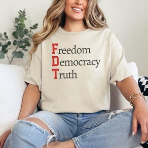 Op de afbeelding: Een persoon draagt een beige T-shirt met de woorden "Vrijheid, Democratie, Waarheid" in rode en zwarte letters.