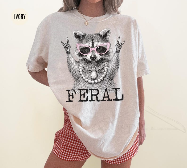 May include: Ivory-colored t-shirt featuring a raccoon wearing pink sunglasses and a pearl necklace. The raccoon is making a rock-and-roll hand gesture. The word "FERAL" is printed in bold, black letters below the raccoon.