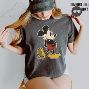 May include: Dark grey Comfort Colors boxy tee featuring a vintage Mickey Mouse graphic. The cartoon character is depicted in his classic pose, with red shorts, yellow shoes, and a smiling expression. The shirt has the text "COMFORT COLORS" and "BOXY TEE".