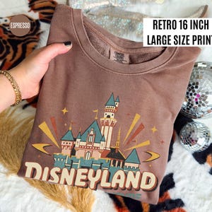 Puede incluir: Sudadera color espresso con un gr&aacute;fico retro del castillo de Disneyland. El gr&aacute;fico es una impresi&oacute;n de gran tama&ntilde;o, de aproximadamente 40,6 cm. La sudadera tiene cuello redondo y mangas largas. El texto "Disneyland" est&aacute; impreso en la parte inferior del gr&aacute;fico.