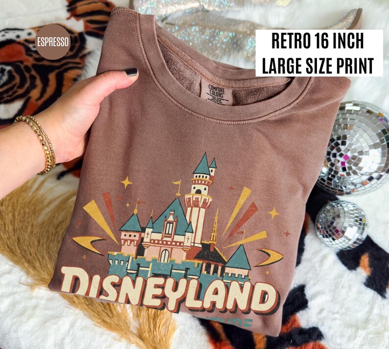 Puede incluir: Sudadera color espresso con un gr&aacute;fico retro del castillo de Disneyland. El gr&aacute;fico es una impresi&oacute;n de gran tama&ntilde;o, de aproximadamente 40,6 cm. La sudadera tiene cuello redondo y mangas largas. El texto "Disneyland" est&aacute; impreso en la parte inferior del gr&aacute;fico.