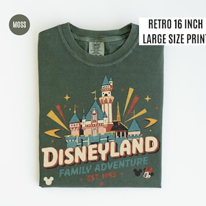 Puede incluir: Una camiseta verde musgo con un gr&aacute;fico retro de Disneyland. El dise&ntilde;o presenta un castillo, la palabra "Disneyland" y el texto "Family Adventure Est. 1955". La camiseta tiene una etiqueta "Retro 16 inch Large Size Print".