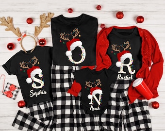 Pode incluir: Pijamas de Natal pretos para a família com chifres de rena, gorros de Pai Natal e letras personalizadas. Os pijamas incluem tops pretos e calças xadrez. Um cardigan vermelho e enfeites de Natal completam a cena festiva.