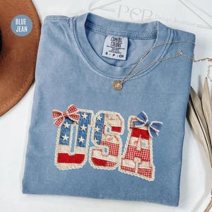 May include: A pale blue t-shirt featuring the word "USA" in a patchwork design. The letters are crafted from fabric with red, white, and blue patterns. A gold necklace with a pendant is draped across the shirt. The shirt is a small size.