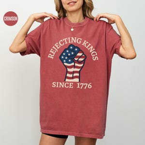Puede incluir: Camiseta de color carmes&iacute; con el texto "Rejecting Kings Since 1776" alrededor de un dise&ntilde;o de pu&ntilde;o. El pu&ntilde;o est&aacute; coloreado con la bandera americana. La camiseta tiene mangas cortas y cuello redondo.