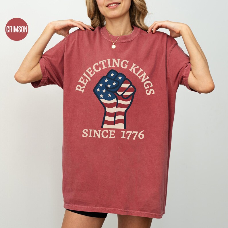 Puede incluir: Camiseta de color carmes&iacute; con el texto "Rejecting Kings Since 1776" alrededor de un dise&ntilde;o de pu&ntilde;o. El pu&ntilde;o est&aacute; coloreado con la bandera americana. La camiseta tiene mangas cortas y cuello redondo.