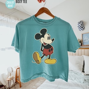 May include: A mint green boxy tee featuring a classic Mickey Mouse graphic. The cartoon character is depicted in black, red, yellow, and beige. The shirt is hanging on a wooden hanger. Text on the shirt reads "COMFORT COLORS CHALKY BOXY TEE MINT".