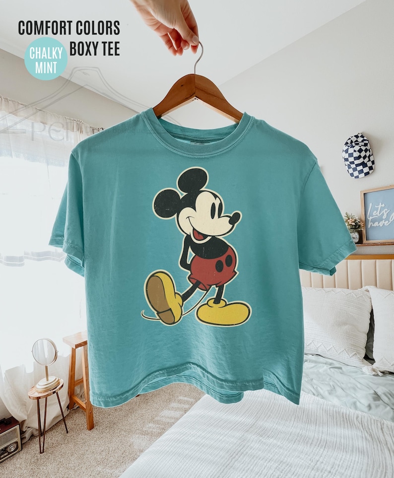 May include: A mint green boxy tee featuring a classic Mickey Mouse graphic. The cartoon character is depicted in black, red, yellow, and beige. The shirt is hanging on a wooden hanger. Text on the shirt reads "COMFORT COLORS CHALKY BOXY TEE MINT".
