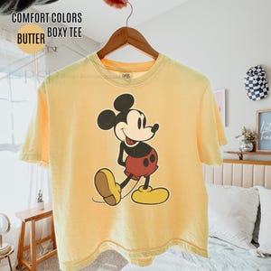 May include: A yellow boxy tee shirt featuring a Mickey Mouse graphic. The shirt is suspended on a wooden hanger. The text on the shirt reads "COMFORT COLORS BUTTER BOXY TEE".