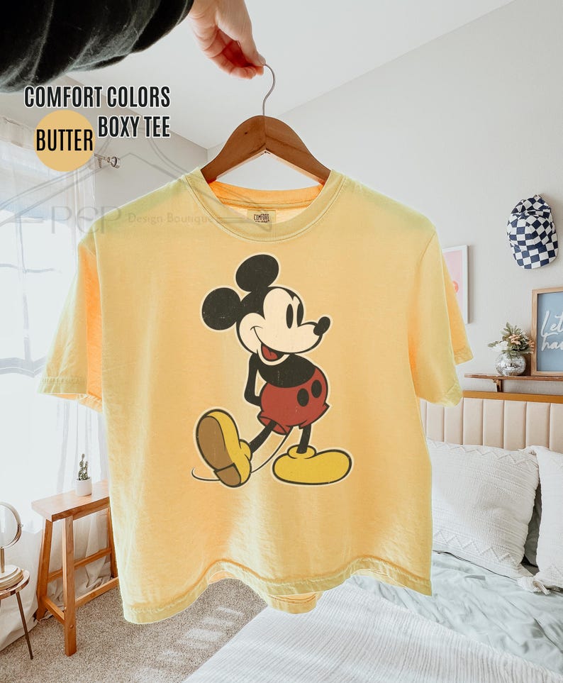 May include: A yellow boxy tee shirt featuring a Mickey Mouse graphic. The shirt is suspended on a wooden hanger. The text on the shirt reads "COMFORT COLORS BUTTER BOXY TEE".